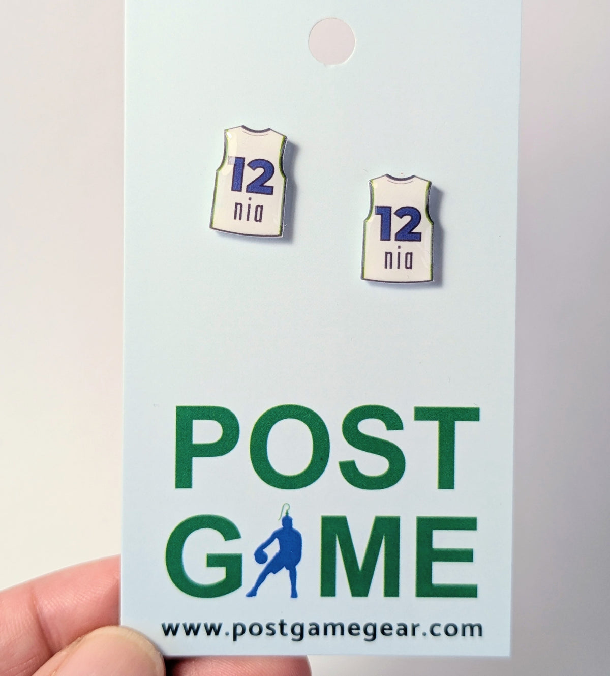 Two small white earrings shaped like basketball jerseys, printed with 12 and nia displayed on a POST GAME branded card held by a hand.