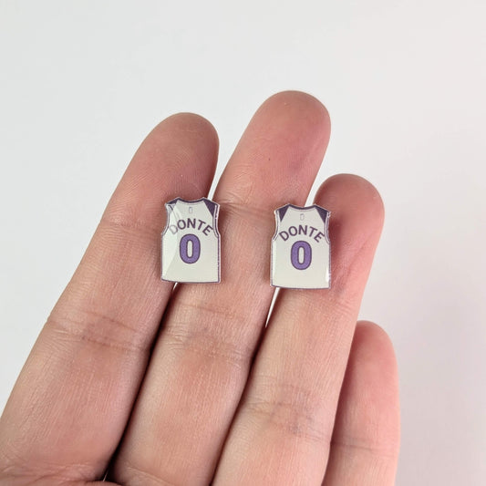 Two small earrings shaped like miniature sports jerseys with 'Donte' and number '0' on a finger against a white background.