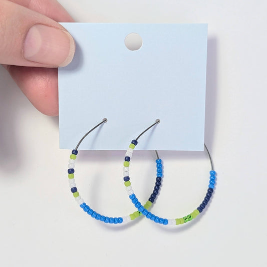 Hoop earrings with blue, green, and white beads on a card held by a hand.