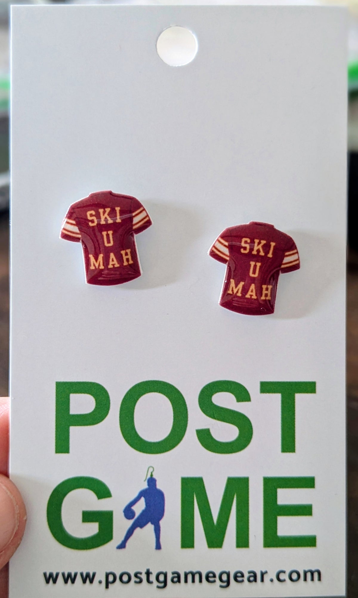 A pair of small earrings shaped like maroon athletic jerseys with yellow text reading 'SKI U MAH', attached to a white display card. The card has large green 'POST GAME' text and a website at the bottom.