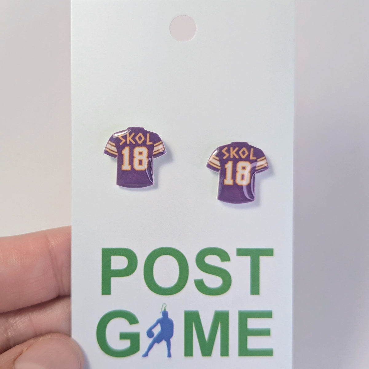 A hand holding a white display card featuring two small earrings shaped like purple Viking football jerseys with the word 'SKOL' and number '18' in gold/yellow. The card also has the green text 'POST GAME'.