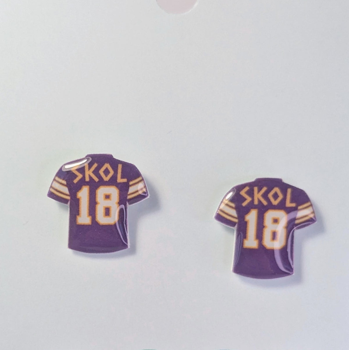 Two small purple sports jersey pins shaped like Vikings football jerseys with the word 'SKOL' and number '18' in gold and white, shown on a plain light background.