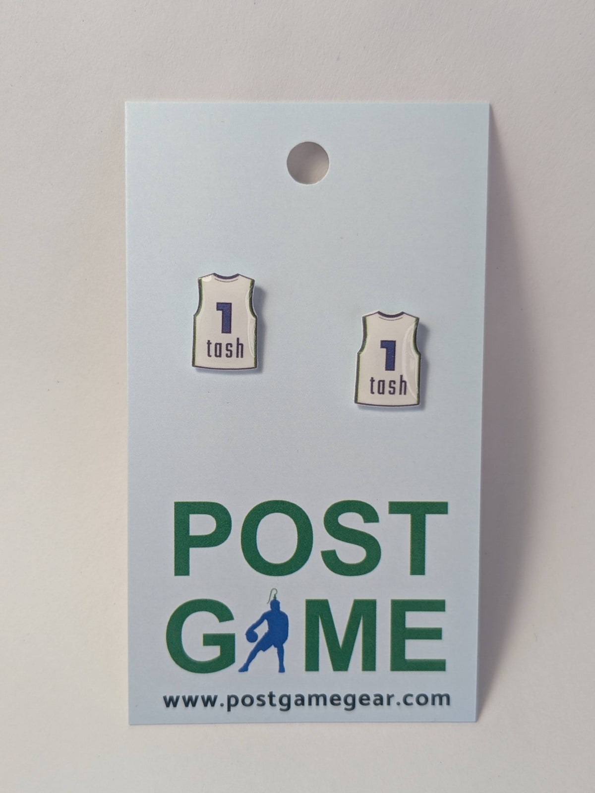 Two earrings shaped like white basketball jerseys with the number 1 and the name 'tosh' in blue, displayed on a light blue Post Game brand card.