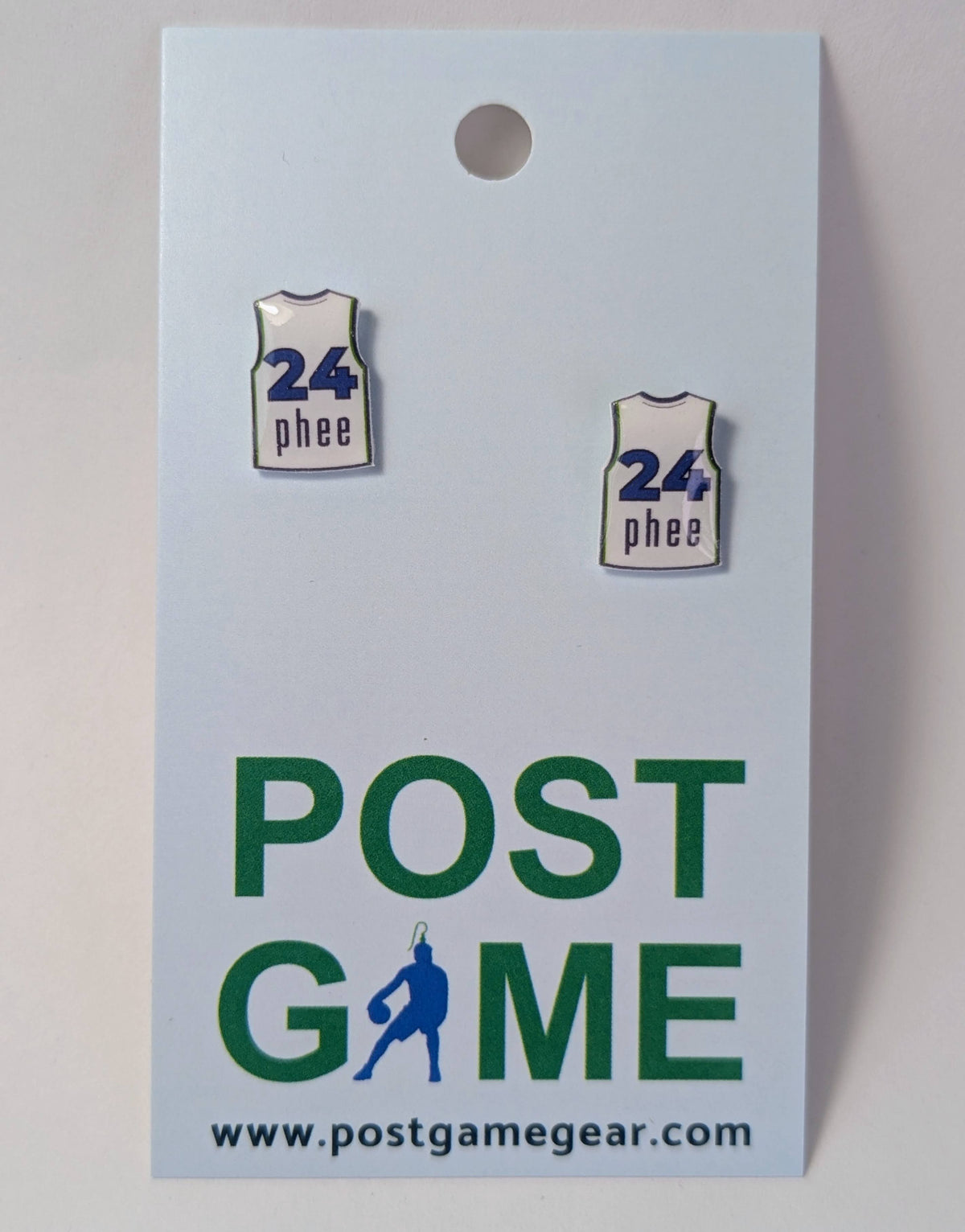 Two small earrings shaped like basketball jerseys with the text '24 phee' in blue on a white background, attached to a light blue display card printed with 'POST GAME' and the website postgamegear.com.