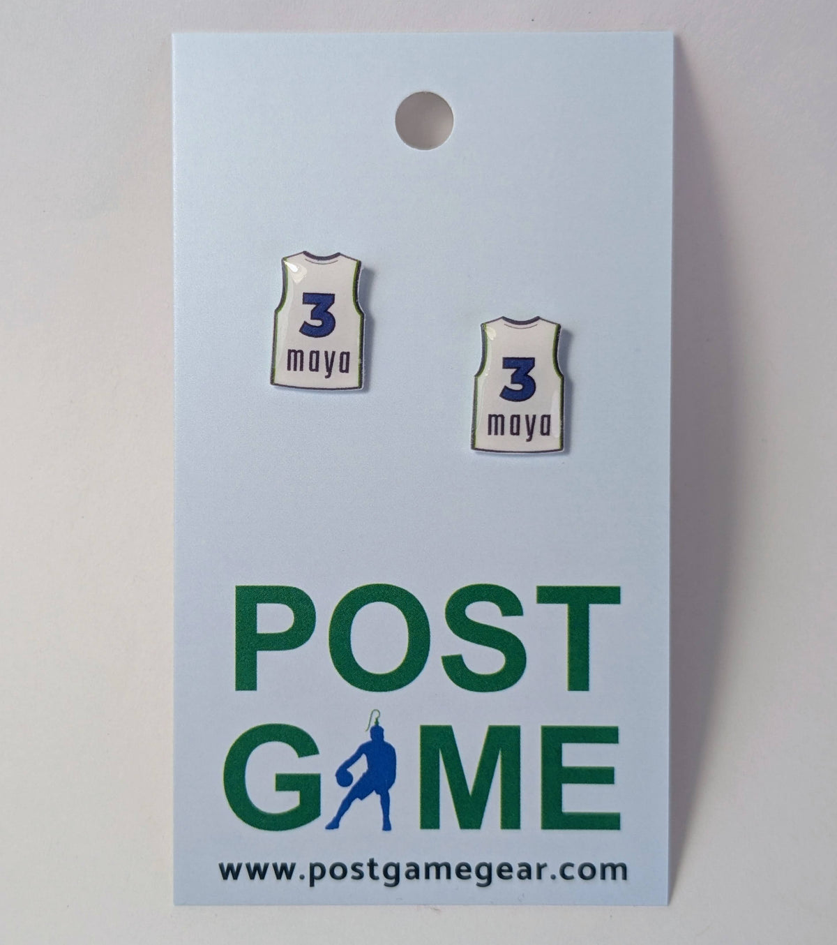 Two small enamel earrings shaped like white basketball jerseys with the name 'maya' and the number '3' in blue, displayed on a light blue backing card printed with 'POST GAME' and the website postgamegear.com.