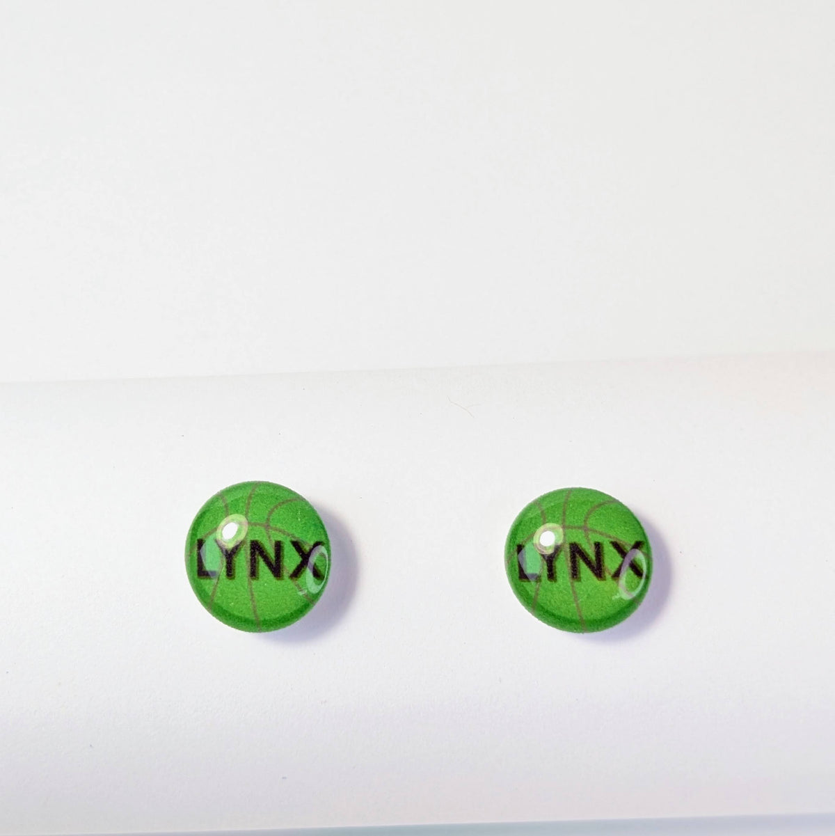 A pair of round green stud earrings with a basketball pattern and the black text 'LYNX' printed on the front, displayed on a clean white surface.
