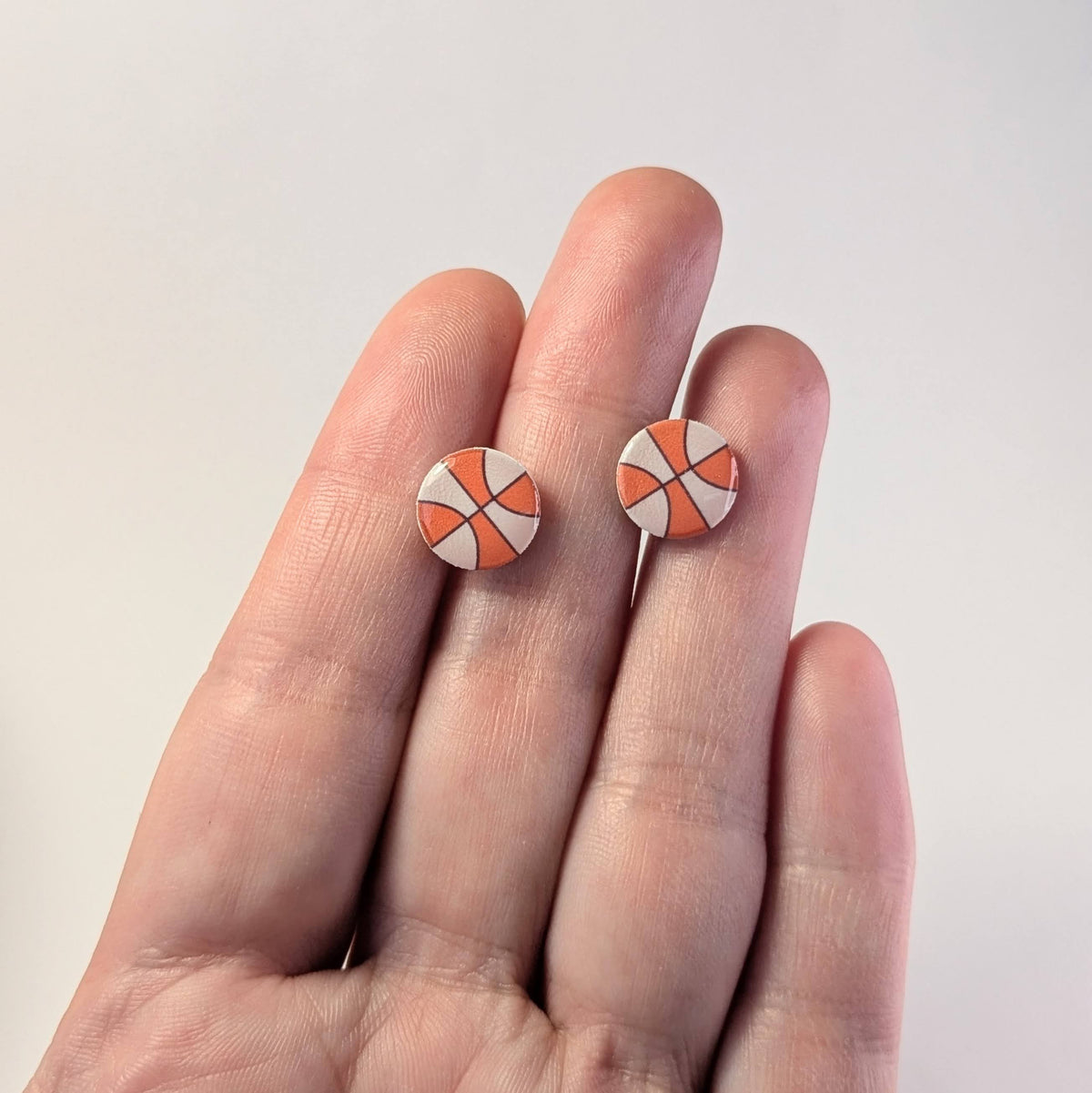 Round earrings with a geometric design like a WNBA basketball, held between fingers on a light background