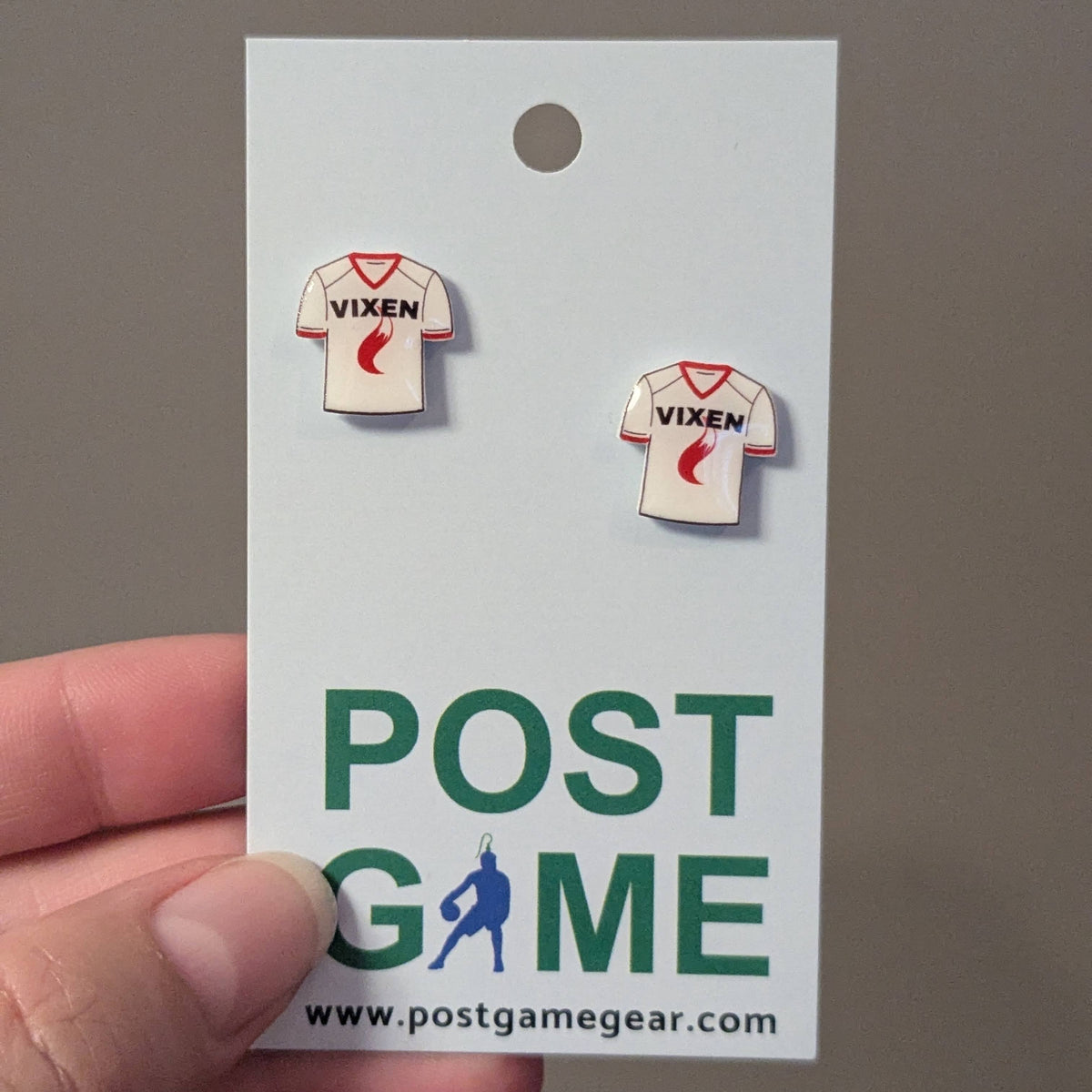 A hand holding a white display card featuring two small enamel earrings shaped like white football jerseys with red trim and the word VIXEN on the chest. The card also has large green and blue text reading POST GAME with the website postgamegear.