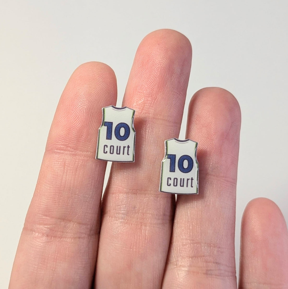 Two small earrings shaped like basketball jerseys with the number 10 and the word 'court' are shown on a person's fingers against a plain light background.