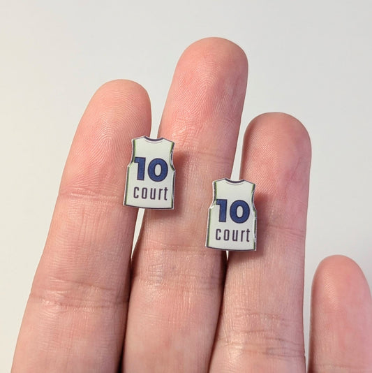 Two small earrings shaped like basketball jerseys with the number 10 and the word 'court' are shown on a person's fingers against a plain light background.