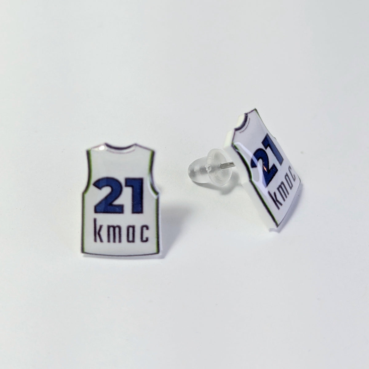 A pair of small earrings shaped like basketball jerseys, shown on a plain white background. One pin is facing forward and displays a white jersey design with the number '21' and the name 'kmac' in dark blue.