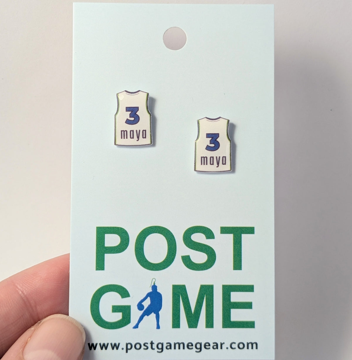 A pair of earrings shaped like white basketball jerseys, printed with 3 and maya, displayed on a card branded with the words POST GAME and held by a hand.