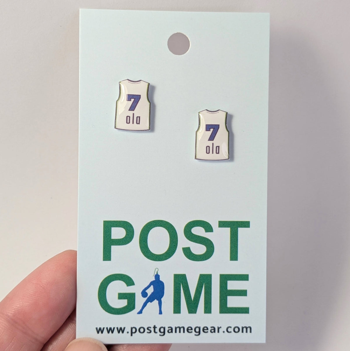 Image of 2 white jersey earrings with the number 7 and name ola on a light colored card, held by a hand. 