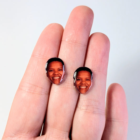 Two oval stud earrings featuring printed portraits of a smiling woman, displayed on a person's fingers against a plain light background.