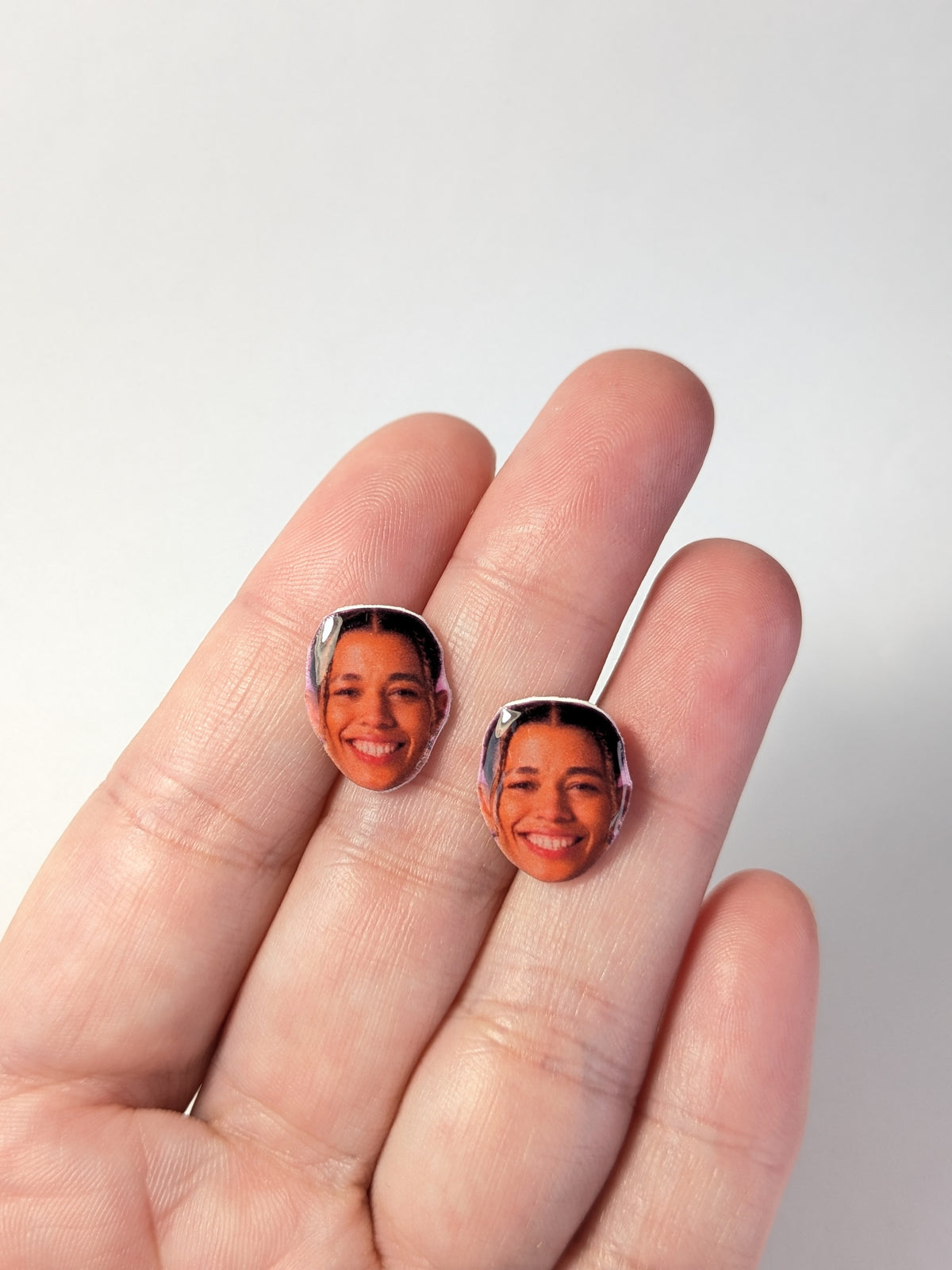 Two small oval stud earrings featuring portraits of a smiliing Natasha Cloud, held on a person's fingers against a plain light background.