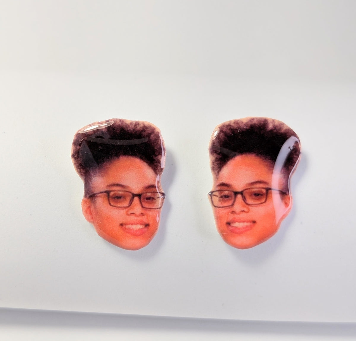 A pair of oval stud earrings featuring printed portraits of a woman with glasses and a high-top hairstyle, displayed on a clean white surface.