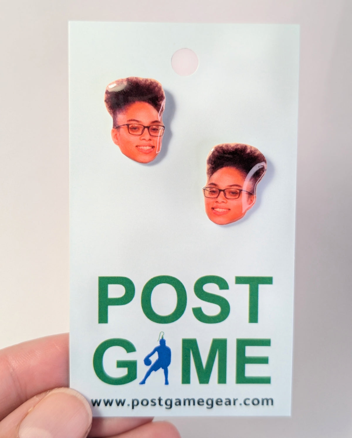 A hand holding a display card featuring two small earrings with printed portraits of a person with glasses and styled hair. The card has green text reading 'POST GAME' and a website at the bottom.