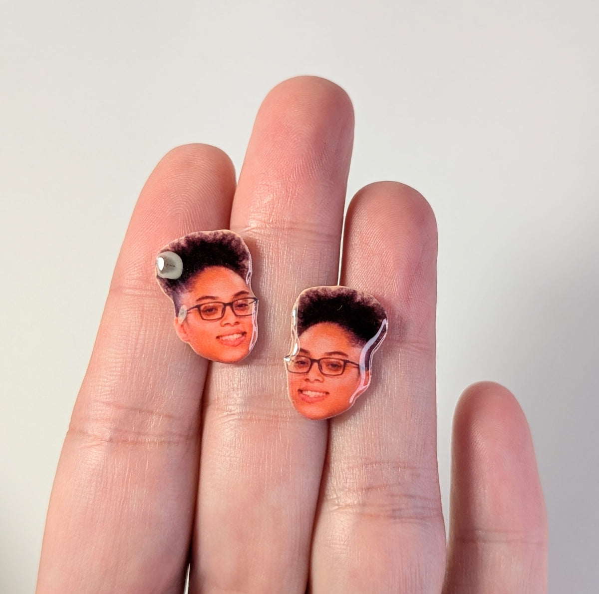 A pair of small portrait stud earrings featuring a woman with glasses and dark hair, shown on a person's fingers against a plain light background.