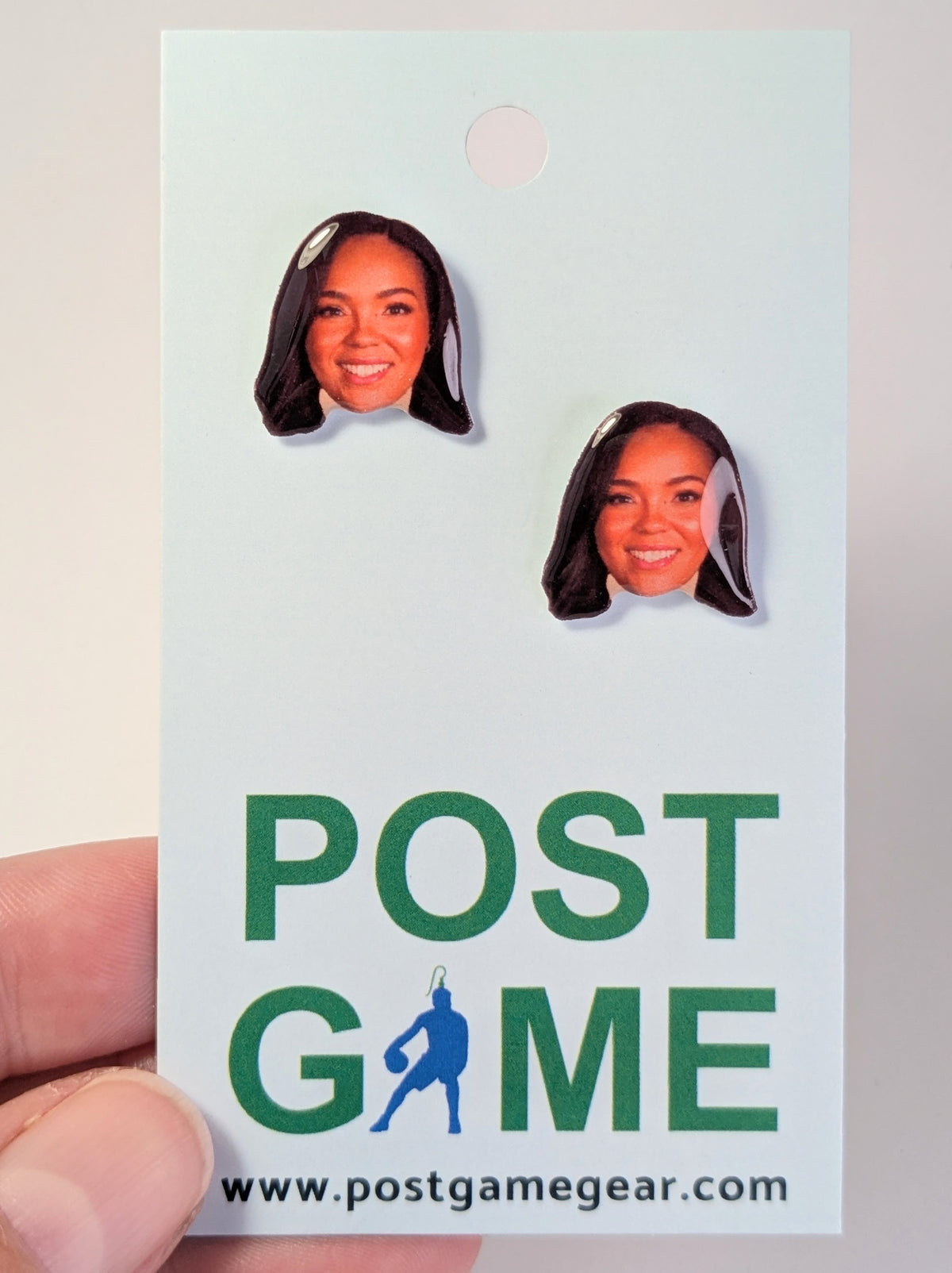 A hand holding a display card featuring two small printed portraits of a woman with dark hair, above the green text 'POST GAME' and the website 'www.postgamegear.