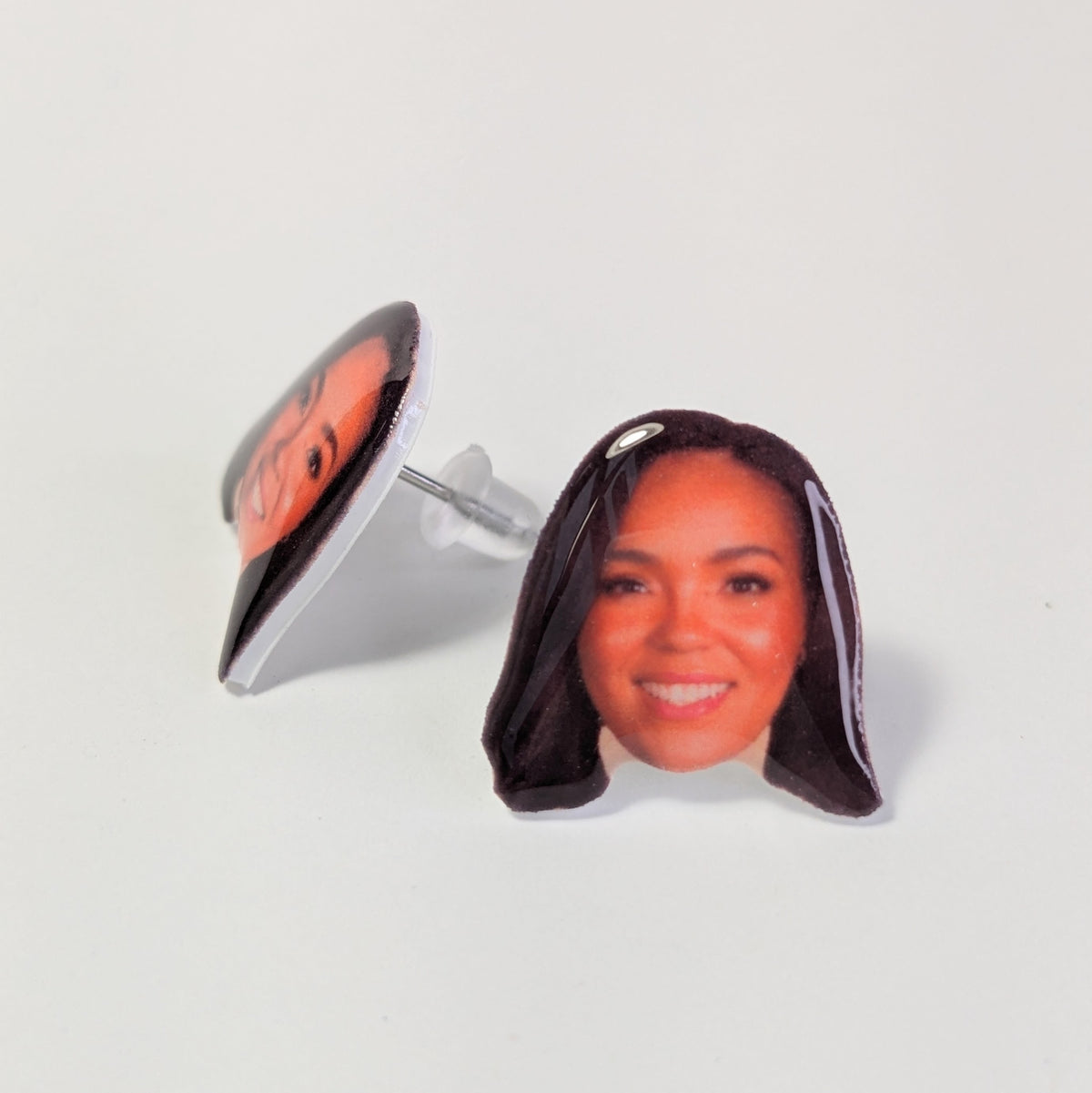 A pair of stud earrings featuring a printed portrait of a smiling woman with dark hair, shown on a clean white background. One earring is shown from the front, and the other is angled to show the back with its clear adhesive backing and metal post.
