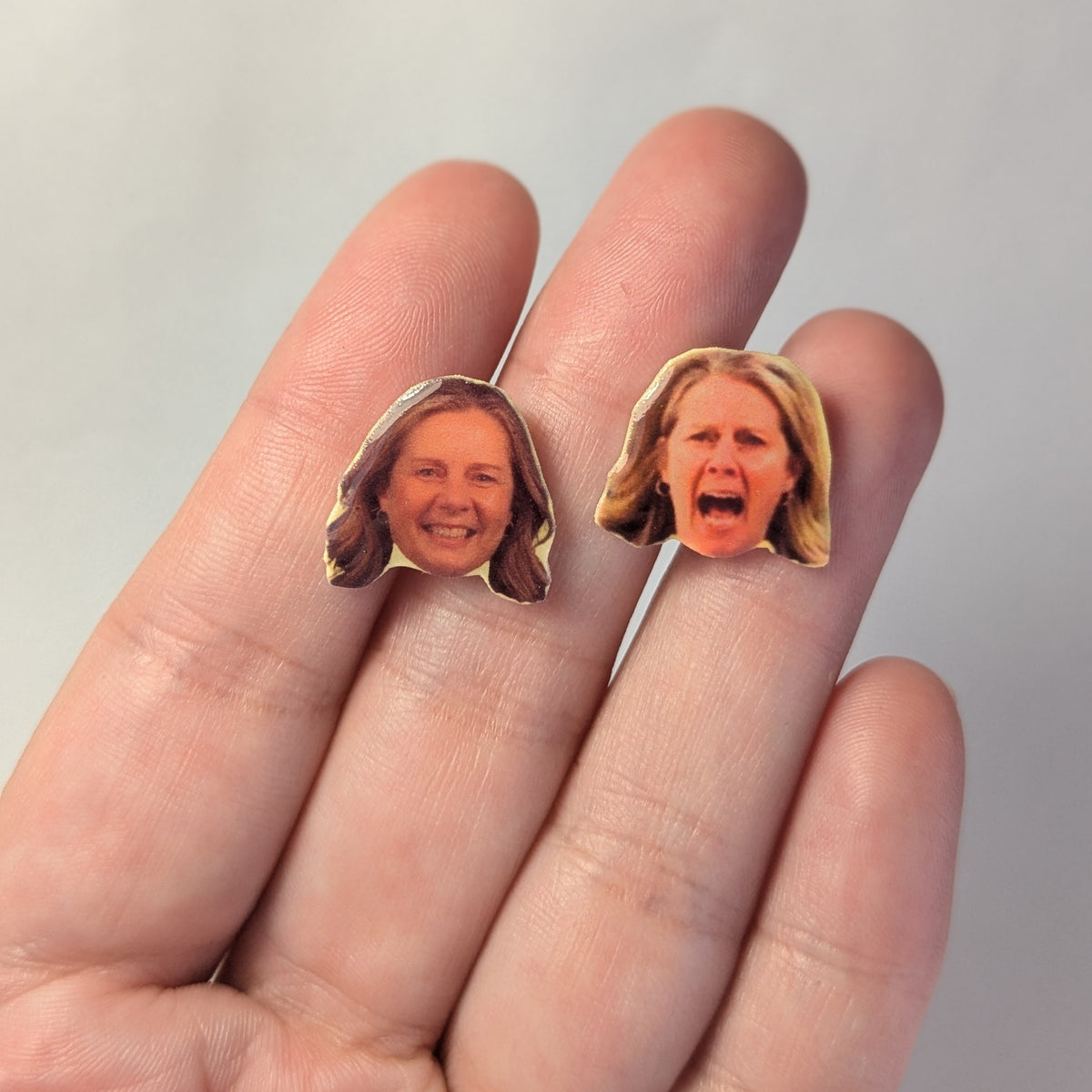 Two small face-shaped earrings displayed on a person's fingers against a plain light background.