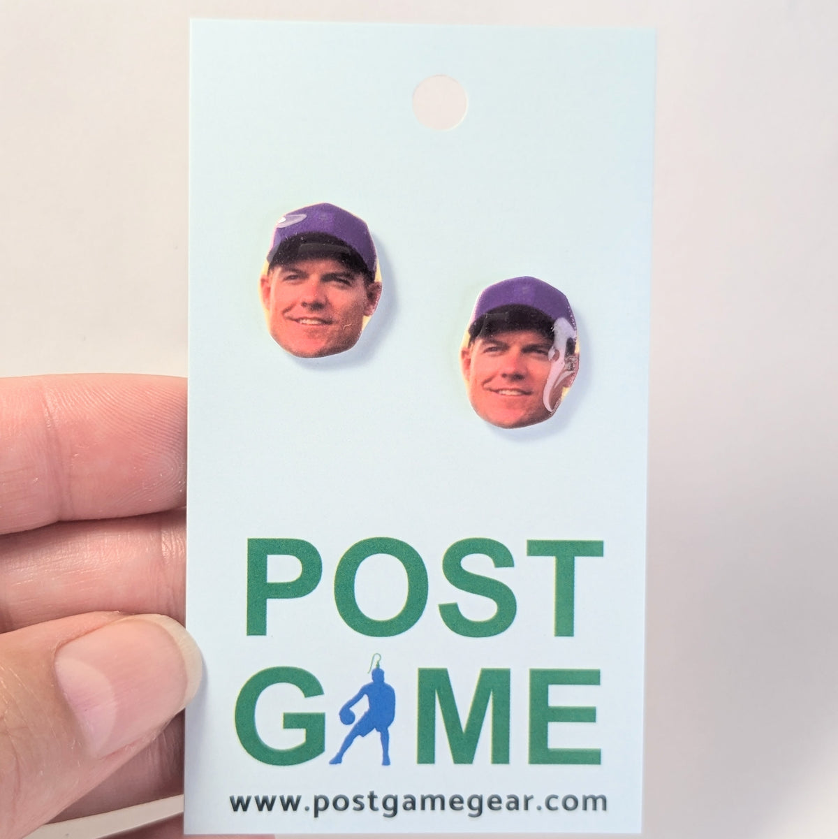 A hand holding a light blue card featuring two earrings with a man's face wearing a purple cap. The card has the text 'POST GAME' in green and blue.