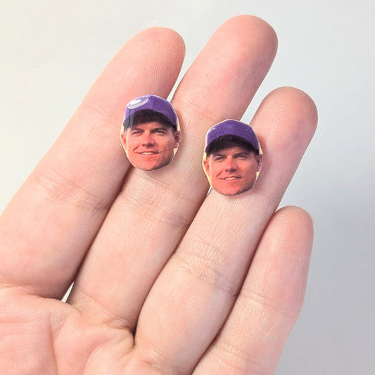 Two small earrings featuring an image of a man in a purple cap, held on a person's fingers against a light gray background.