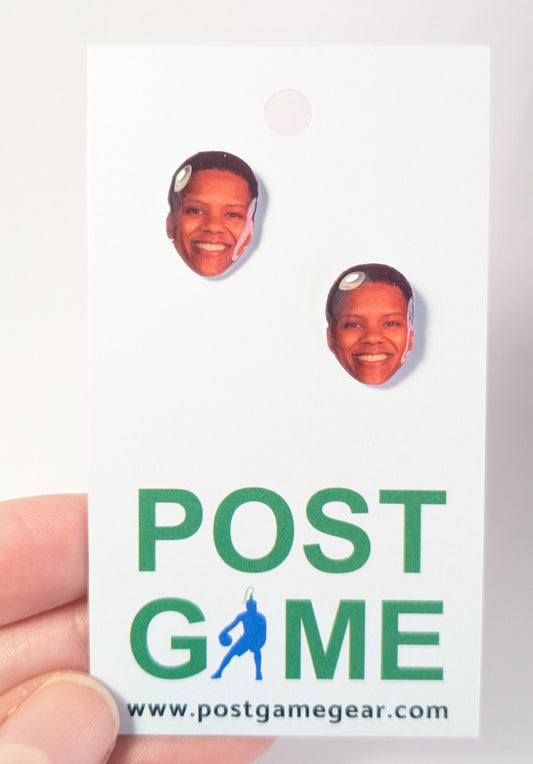 A hand holding a display card featuring two oval-shaped face earrings with a cutout of a smiling face. The card has green text reading 'POST GAME' with a blue silhouette graphic and a website.