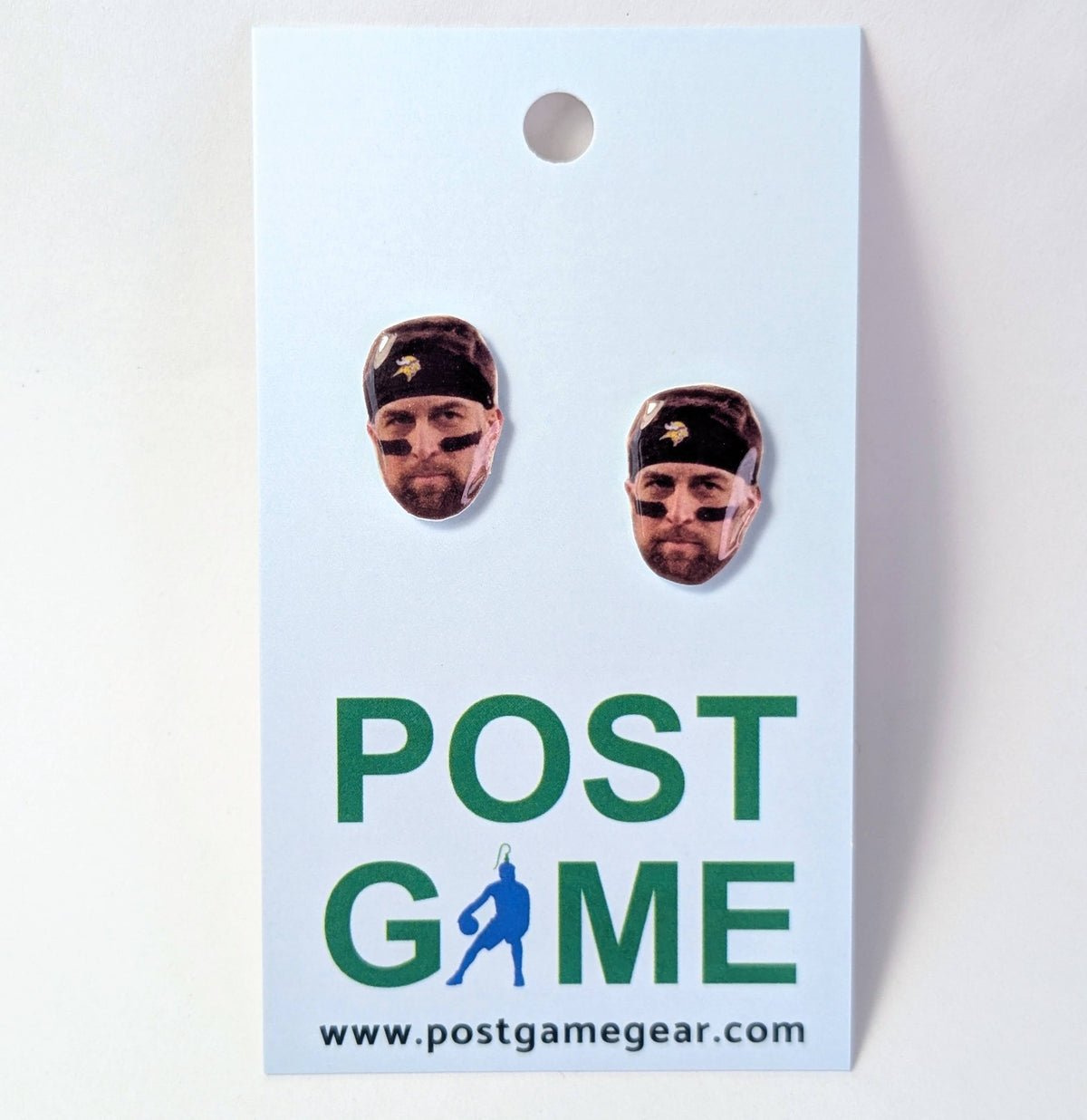 A pair of custom earrings shaped like football player faces with black face paint ,displayed on a white card that reads 'POST GAME' with the website 'www.postgamegear.
