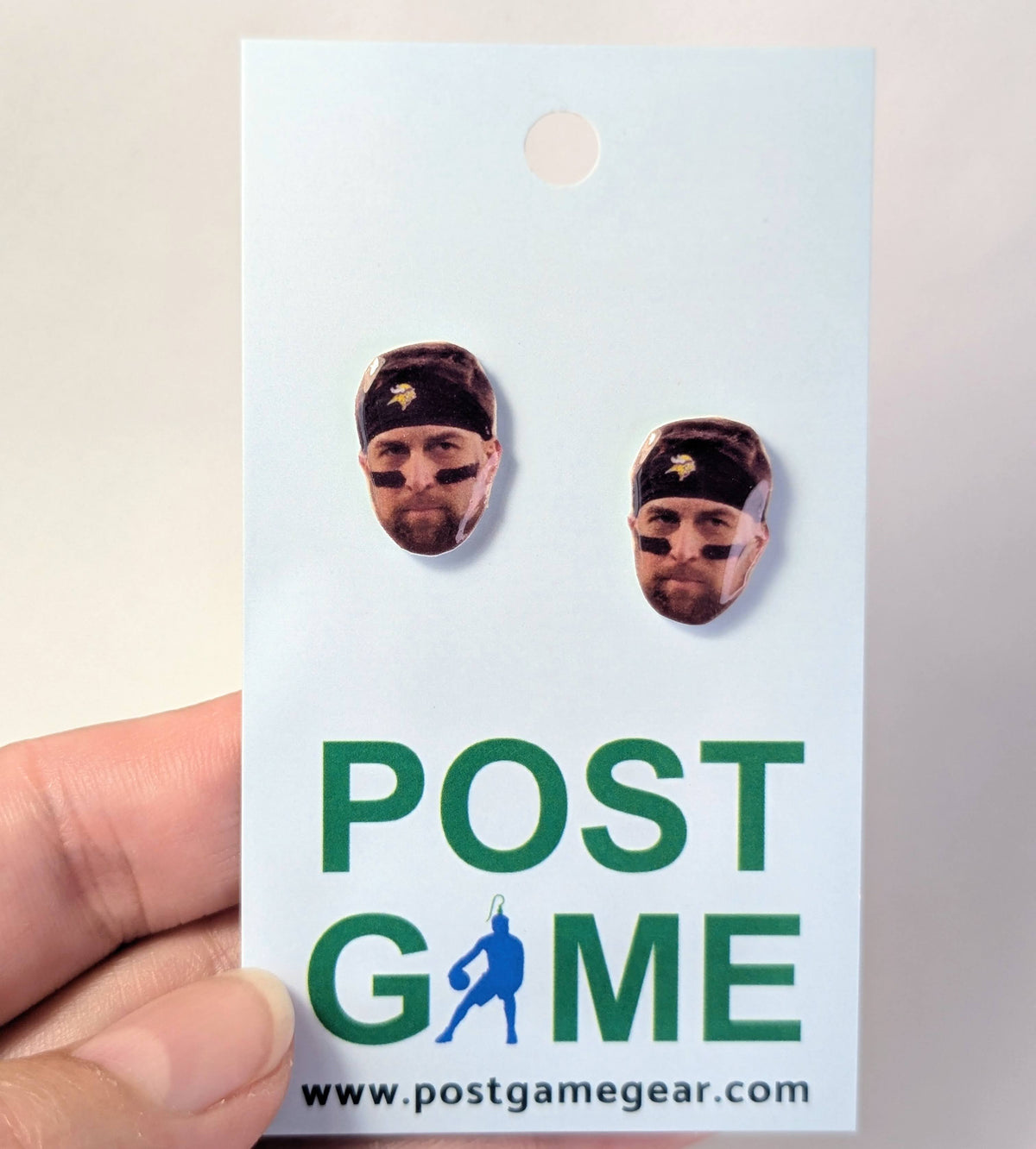 A hand holding a white display card featuring two small oval earrings with football player faces and the text 'POST GAME' and 'www.postgamegear.