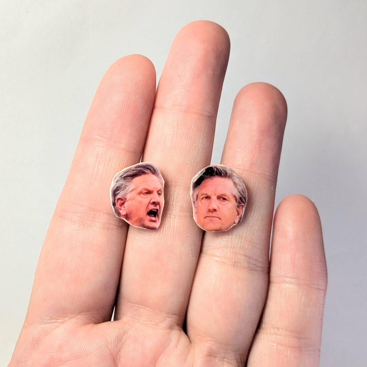 Two small oval earrings with printed faces resting on a person's fingers against a plain light background.