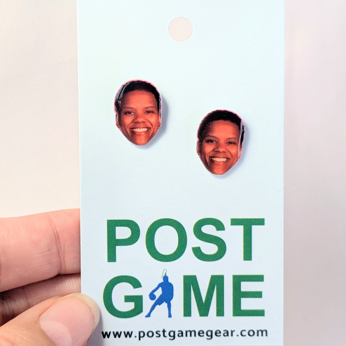 A hand holding a light blue card featuring two oval-shaped face cutouts with printed portraits, above the green text 'POST GAME' and the website 'www.postgamegear.