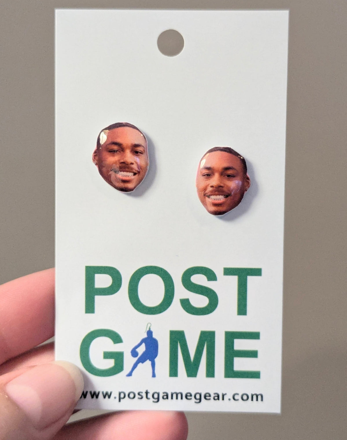 A hand holding a white display card featuring two earrings with a printed photo of a smiling man, above the green text 'POST GAME' and the website 'www.postgamegear.