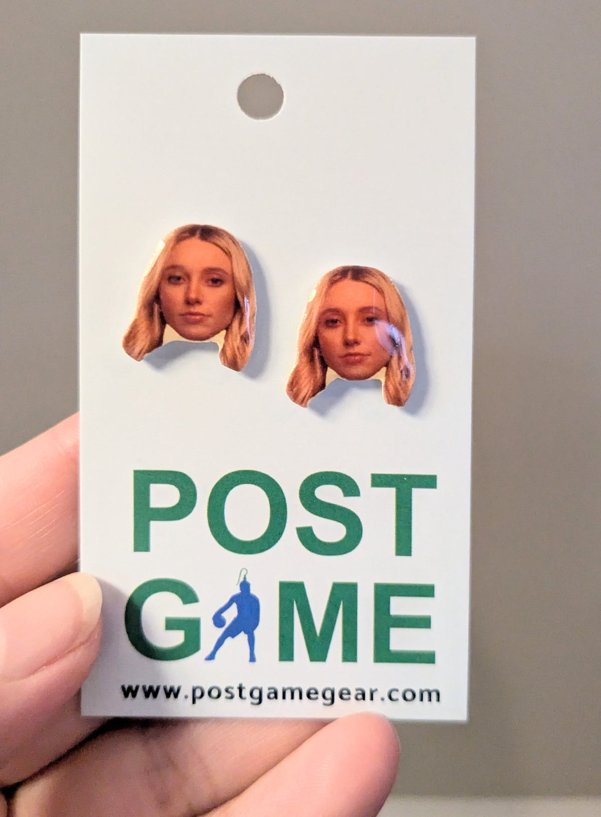 A hand holding a white display card featuring two small earrings with cutout portraits of a blonde woman, above large green text reading POST GAME and a website URL. 