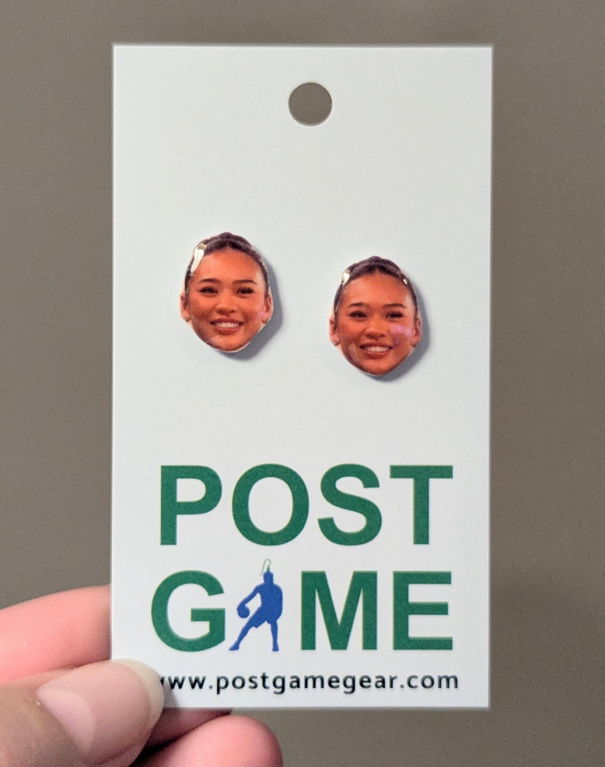 A hand holding a white display card featuring two oval stud earrings with printed faces of a smiling woman. The card has green text reading 'POST GAME' and a website at the bottom.