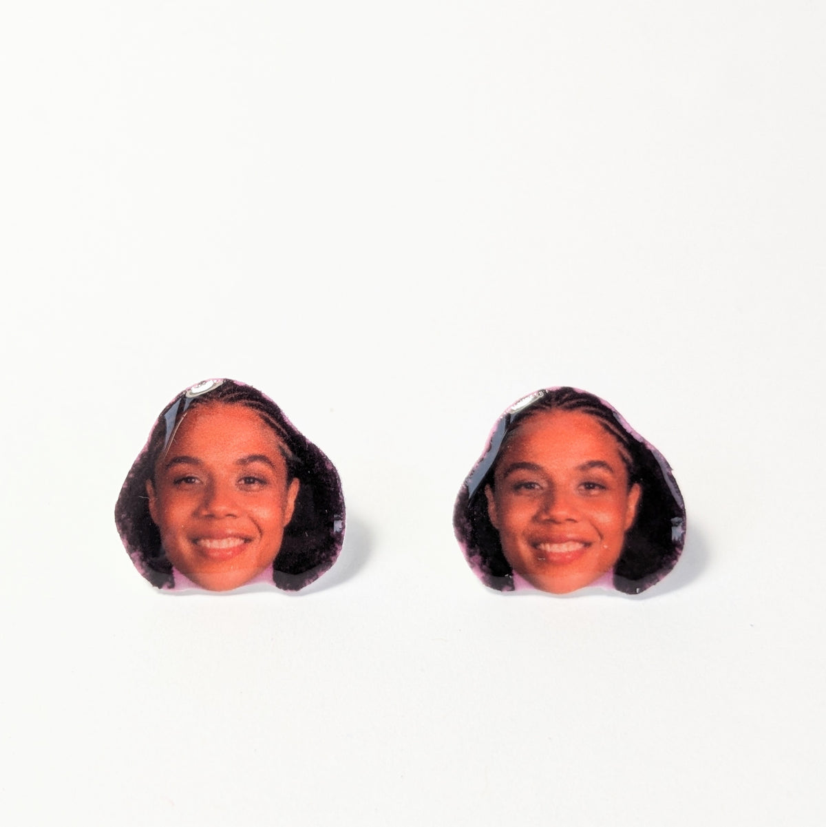 A pair of novelty stud earrings shaped like the head of a woman (Nia Brodie) with dark hair, shown on a plain white background.