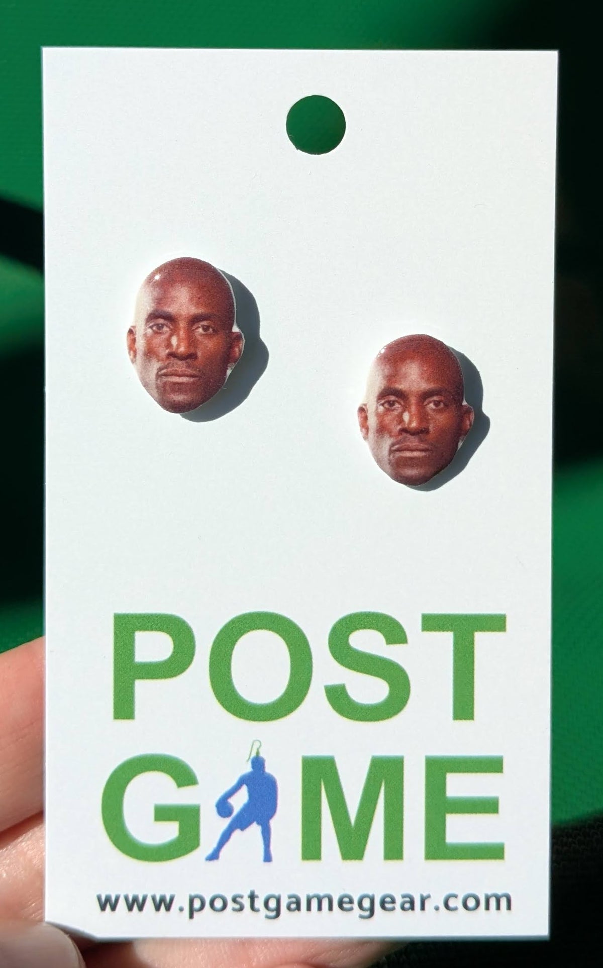 A pair of novelty earrings shaped like the face of Kevin Garnett, mounted on a white display card printed with 'POST GAME' and a website, held against a dark green background.