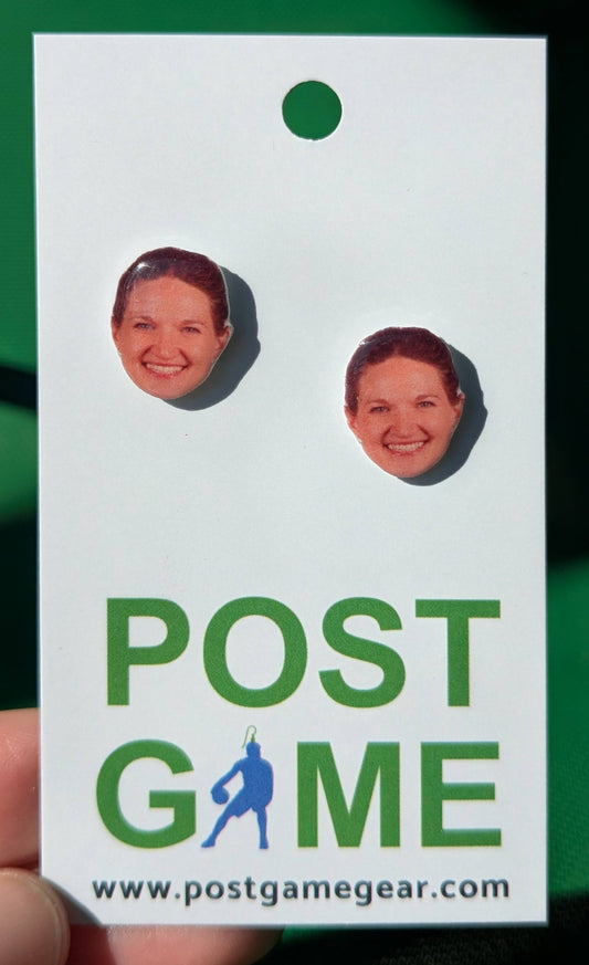 A pair of stud earrings shaped like the face of Lee Stecklein, attached to a white display card printed with the words 'POST GAME' and a website, held against a dark green background.