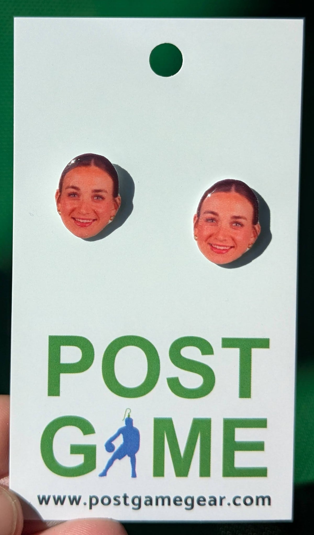A white post game merchandise card featuring two earrings cutouts of Taylor Heise's face with green text 'POST GAME' and the website 'www.postgamegear.