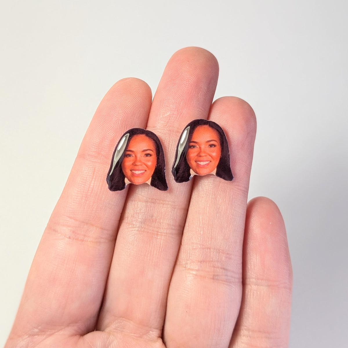 A pair of miniature face stud earrings featuring a portrait of a woman with dark hair, held on a person's fingers against a plain light gray background.