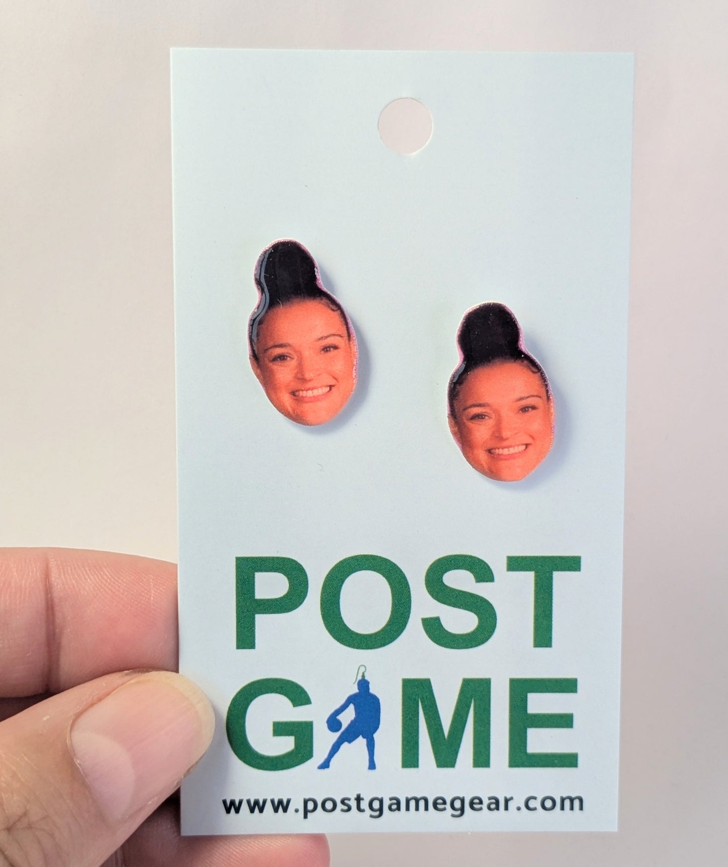 A hand holding a white display card featuring two portrait earrings, with the text 'POST GAME'.