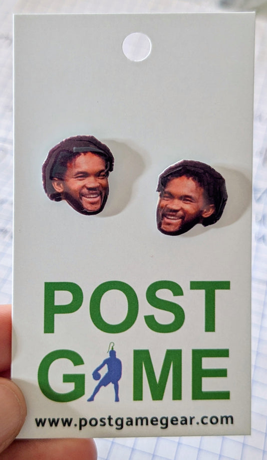 A pair of novelty stud earrings featuring printed images of Kyler Murray displayed on a white backing card with green text reading 'POST GAME' and a website URL. A hand is holding the card.