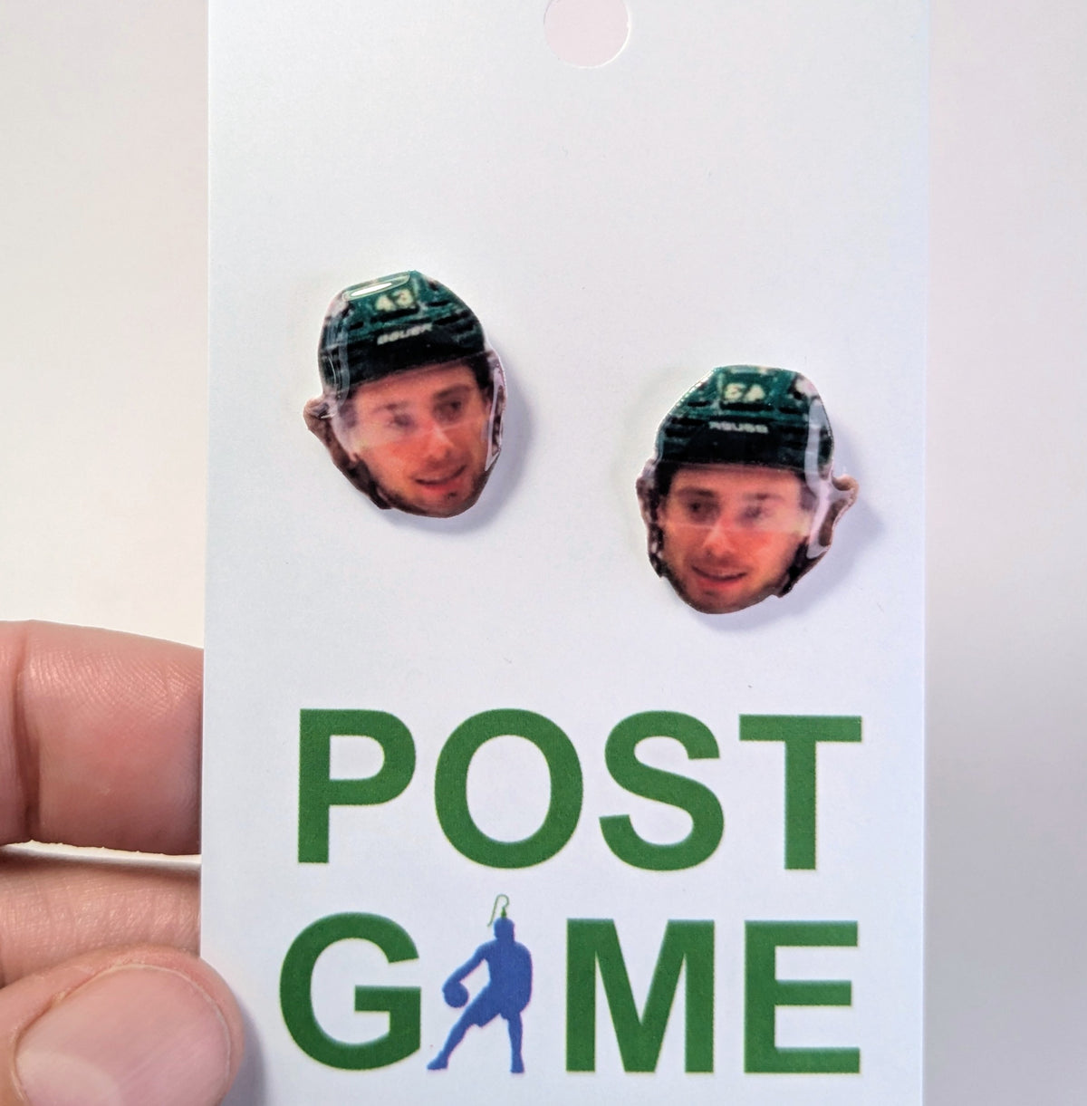 A hand holding a white card featuring two faces of hockey player Quinn Hughes in a green helmet, with the text 'POST GAME' printed below in green and blue.