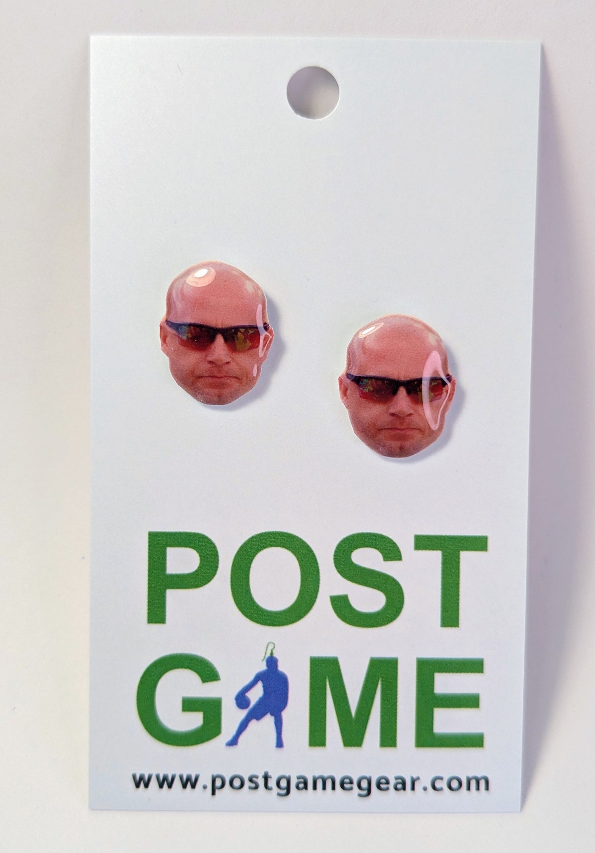 A pair of oval stud earrings featuring printed bald men's face wearing sunglasses, mounted on a white display card with the text 'POST GAME' and 'www.postgamegear.
