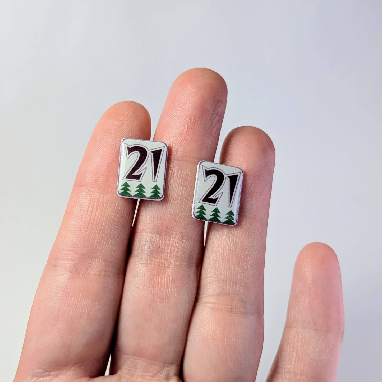 Two small square earrings with '21' design held between fingers against a light gray background