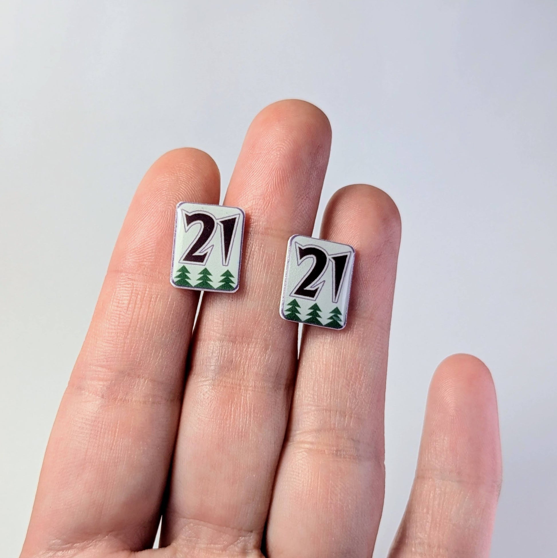 Two small square earrings with '21' design held between fingers against a light gray background