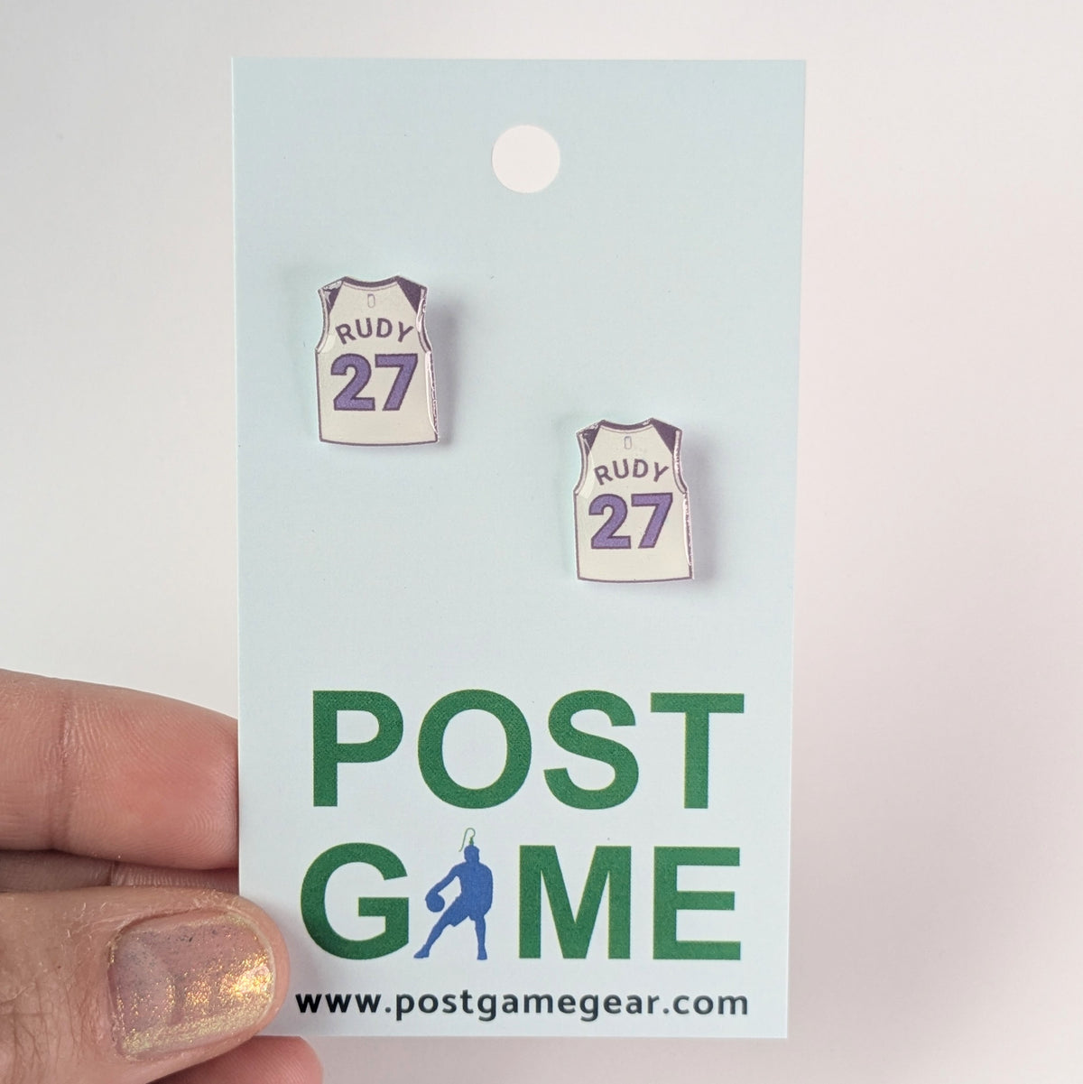 Pair of earrings shaped like a basketball jersey with 'Rudy 27' on a 'Post Game' branded card.