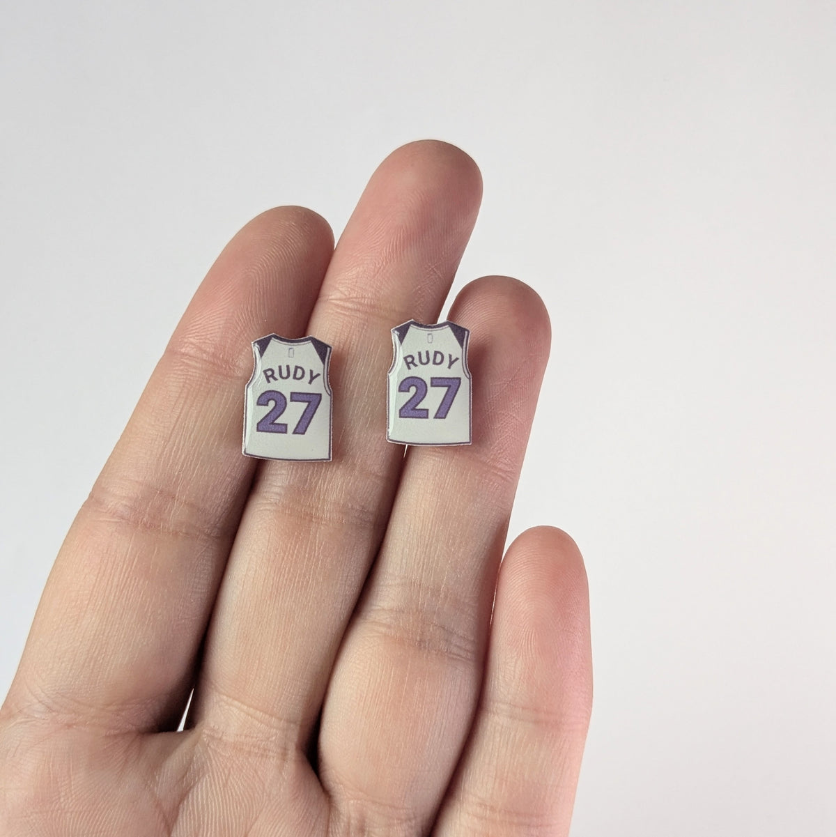 Two earrings shaped like white basketball jerseys with 'RUDY' and number '27' on a hand against a light background.