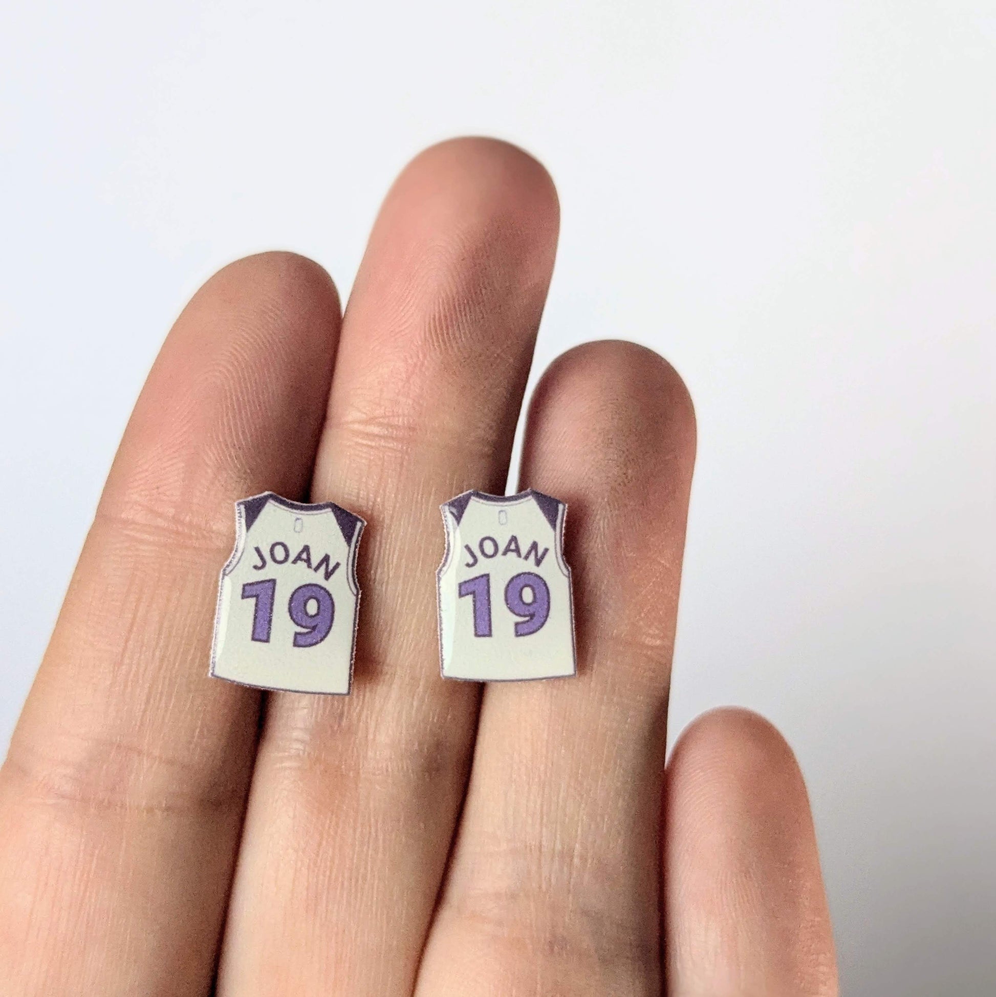 Earrings shaped like miniature basketball jerseys with 'JOAN 19' text on a hand against a white background.
