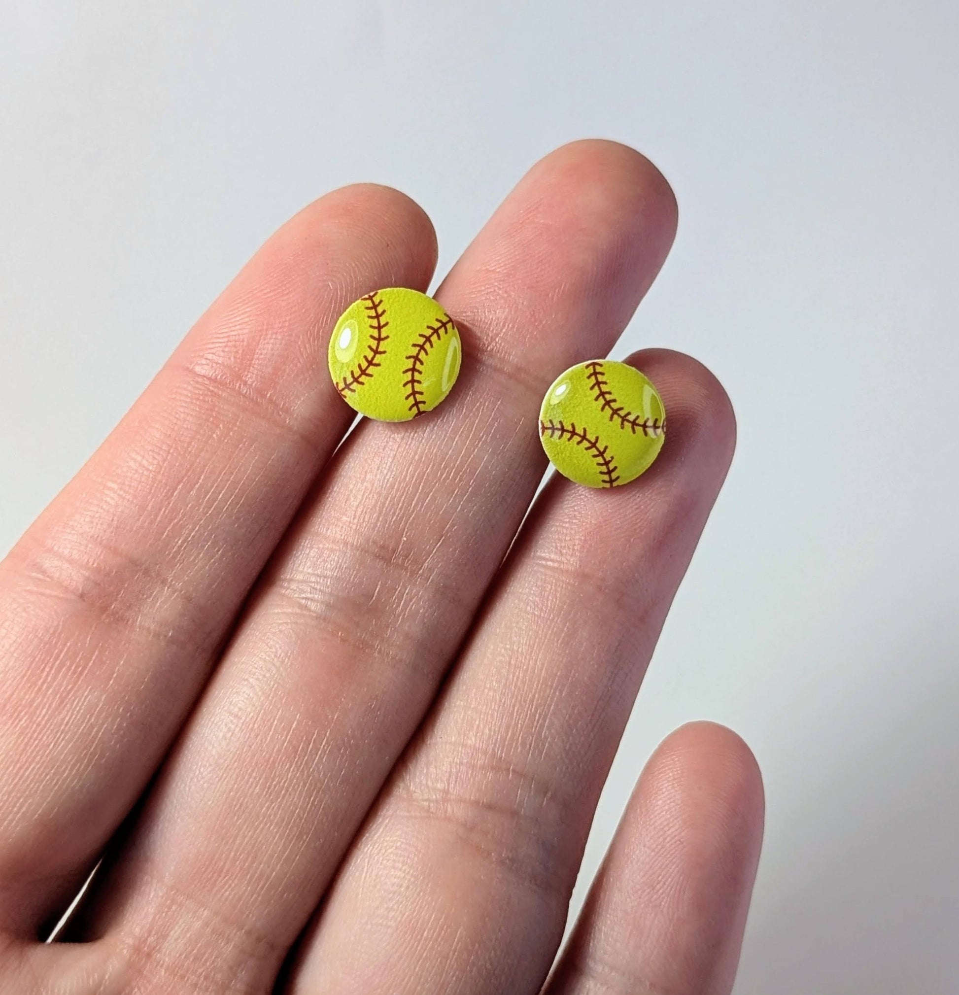 Yellow softball-shaped earrings on a hand against a light background